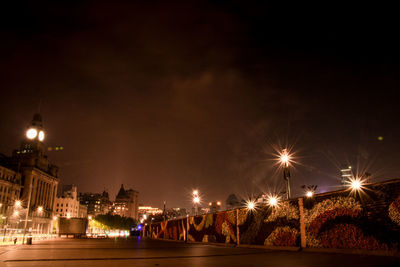 Illuminated street by buildings against sky at night