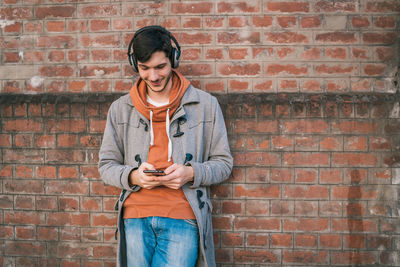 Full length of a young man using mobile phone against brick wall