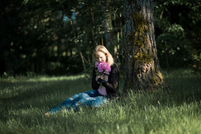 Young woman smelling flowers while sitting on grassy field in forest