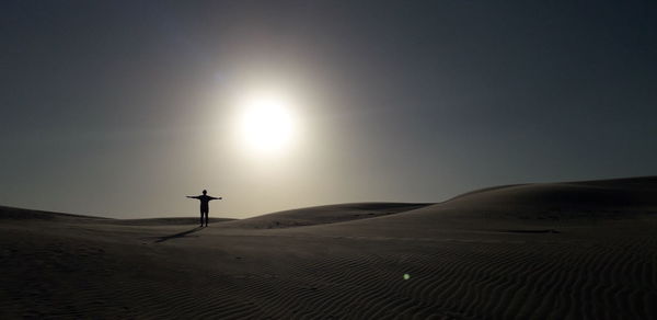 Silhouette man with arms outstretched standing on sand in desert against sky