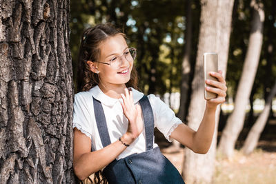 Cheerful pupil in glasses makes a video call on a smartphone near a tree in the park