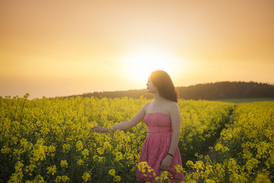 Woman standing amidst yellow flowering plants on field against sky during sunset