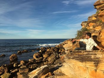 Young man sitting on rock at beach against blue sky