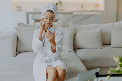 Young woman applying moisturizer while looking at mobile phone at home