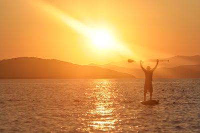 Silhouette man paddleboarding in sea against orange sky