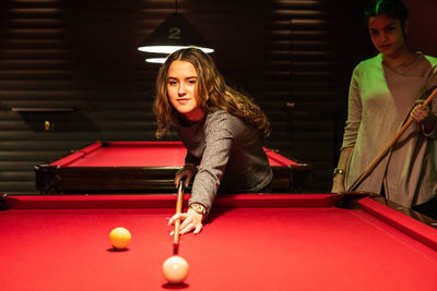 Smiling teenage girl aiming cue ball on illuminated red pool table by friend