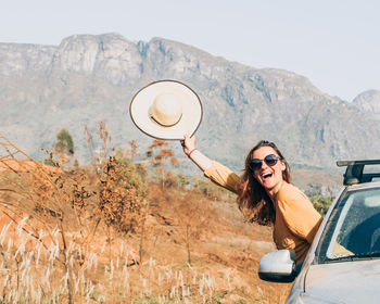 Woman with hat in hand with body getting out of the car with serra do caraça in the background.