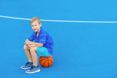 Portrait of boy sitting on basketball at court