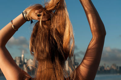 Rear view of woman tying blond hair at beach against sky during sunset