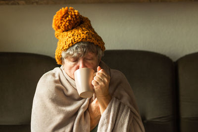 Elderly woman in ginger cap wrapped in blanket drinking cup of tea. old grandma feeling cold at home