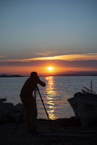 Silhouette man photographing at sea against sky during sunset