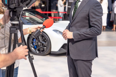 Media interview with car salesperson, sports car in the background