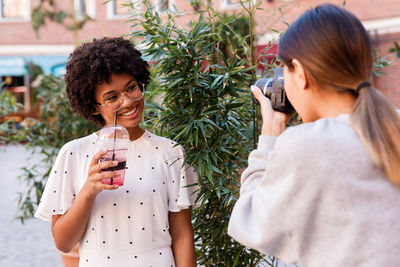 Teenage girl photographing friend holding drink while standing outdoors
