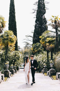 Happy lovers the bride and groom in wedding outfits walk among plants and palm trees in the old park