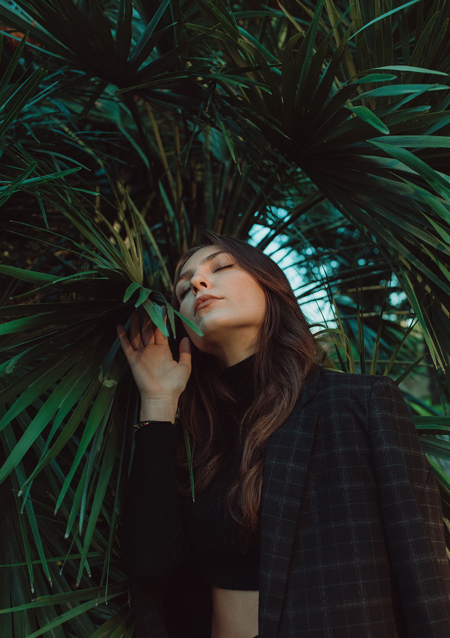 one person, young adult, plant, green, adult, women, nature, long hair, tree, leaf, standing, palm tree, hairstyle, leisure activity, lifestyles, clothing, portrait, looking, plant part, outdoors, casual clothing, tropical climate, front view, brown hair, growth, person, beauty in nature, three quarter length, sunlight, eyes closed, jungle, palm leaf, waist up, darkness, human face, emotion