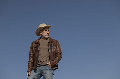 Portrait of adult man with brown leather jacket and cowboy hat against blue clear sky