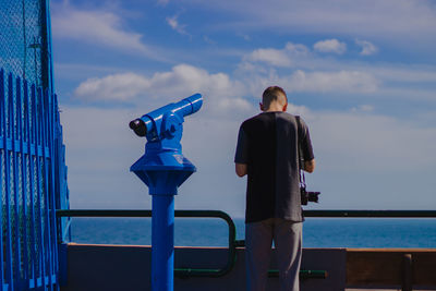 Rear view of man standing by coin-operated binoculars at railing against sea at observation point