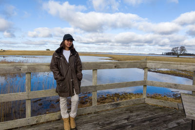 Full length portrait of young woman standing on railing against sky