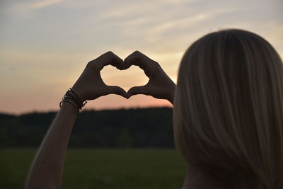 Midsection of woman making heart shape against sky during sunset