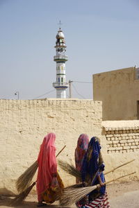3 women in traditional clothing, can be seen carrying brooms with a mosque in the background.