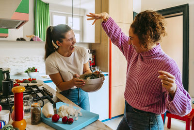 Playful young female friends enjoying while making salad in kitchen at home