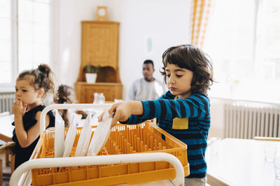 Boy placing plate in crate on cart by girl in classroom at kindergarten