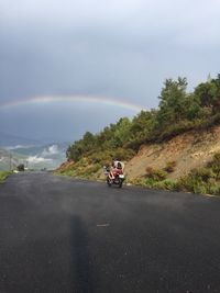 People riding motorcycle on road against rainbow