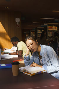 Confident female student reading book while sitting at desk in university library
