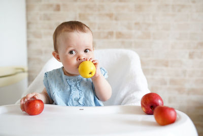 Portrait of cute baby girl holding apples while sitting on high chair at home