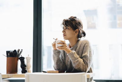 Thoughtful woman looking away while sitting with coffee cup at table in cafeteria