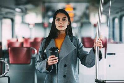 A happy brunette with coffee in her hands stands on the train, looks into the distance and smiles