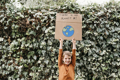 Happy girl with arms raised holding banner with message and planet drawing in front of plant