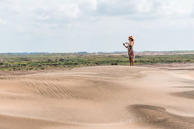 Mature woman using the mobile to take a photo in a desert with plants on a windy day.