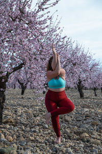 Unrecognizable curvy female in sportswear standing in garudasana in garden with blossoming almond trees and practicing yoga while balancing on leg