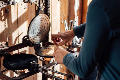 Cropped unrecognizable artisan using stick while rolling up hot freshly baked catalan neula waffles on waffle iron placed over gas flame