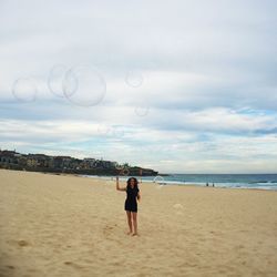 Young woman making bubbles at beach against cloudy sky