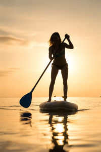 Back view of silhouette of unrecognizable female surfer standing on paddleboard and rowing against spectacular sun in sunset sky