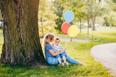 Young caucasian mother and boy toddler son sitting together in park with balloons. 