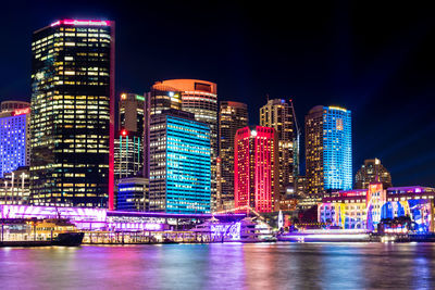 Illuminated buildings by river against sky at night