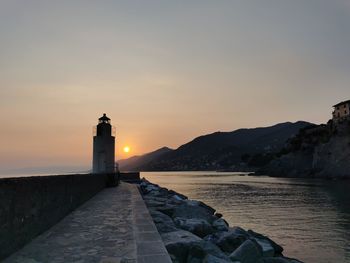 Lighthouse amidst sea and buildings against sky during sunset