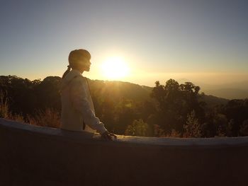 Side view of woman standing by retaining wall on mountain against sky during sunset