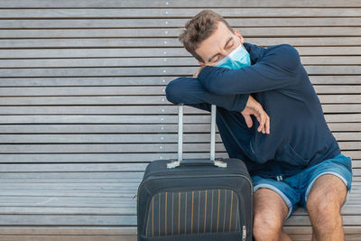 Man wearing flu mask sitting with suitcase outdoors