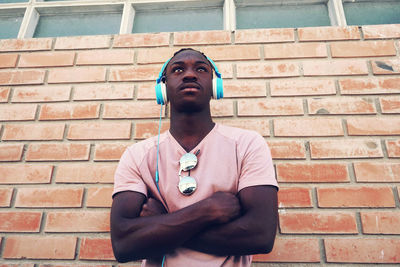 Low angle view of teenage boy with arms crossed listening music while standing against brick wall