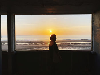 Rear view of woman standing at beach against sky during sunset