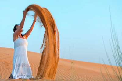 Woman playing with sands at desert against clear sky