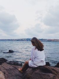 Rear view of woman sitting on rock by sea against sky