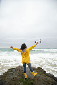 Carefree young woman holding megaphone while standing against sea