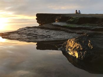 Friends sitting on rock formation against sea during sunset