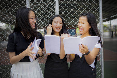 Smiling friends holding book while standing against chainlink fence