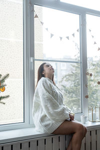 Woman in warm white winter sweater sitting on the window at home at christmas eve holding cup